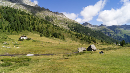 Fototapeta na wymiar Panorama montano con baita, ruscello, alberi e turisti che riposano