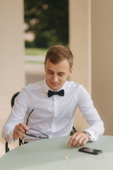 Handsome man in shirt and bow tie use phone. blonde hair man outside. Man sist by the table and try on sunglasses