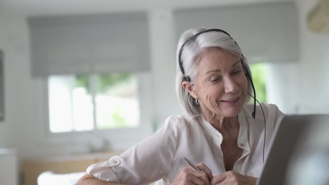 Senior woman talking on headset infront of laptop at home