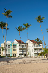 Photo of multy-family coast houses near Atlantic Ocean on beach of Bavaro. Lots of palm trees are planted near house. Roofs of the buildings are darker than facades and are made of terracotta tiles.