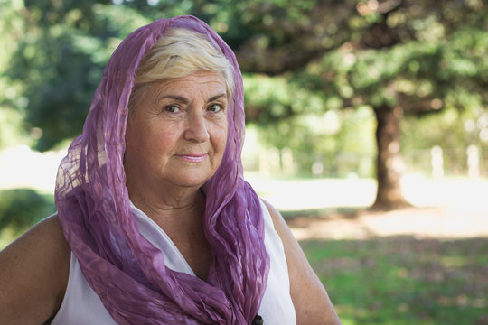 Portrait of young looking woman covering her head with hijab style purple shawl in the park. Mature lady with veil over head, Muslim, Arab look. Female model with charming, serene expression