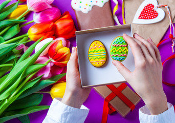 Female hands are holding a gift box with Easter cookie eggs near tulip flowes before wrapping on...