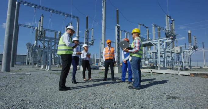 Medium shot of a group of electrical workers in safety vests talking to each other against the background of a transformer station