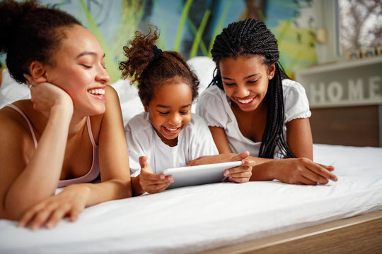 Family time-Smiling girl playing with mother and sister at home in a bed on digital tablet