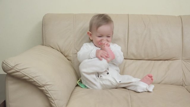Happy baby sits on sofa and plays with his foot laughing
