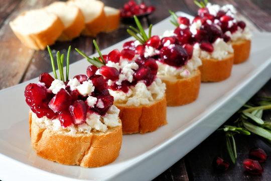 Holiday crostini appetizers with cranberries, pomegranates and feta cheese. Close up on a serving plate against a wood background.