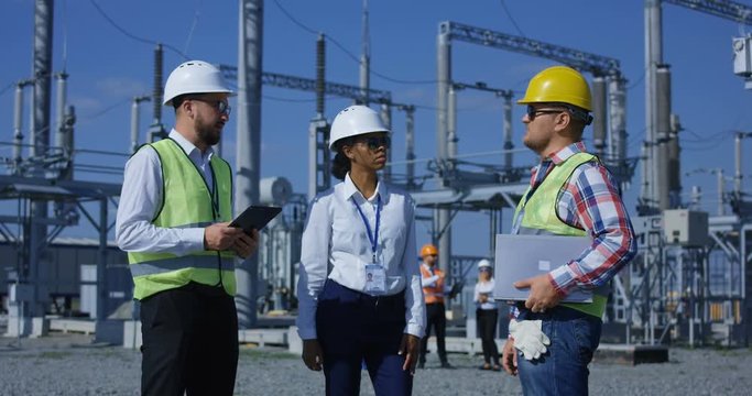 Medium shot of three electrical workers reviewing documents on a tablet during an inspection against the background of a transformer station