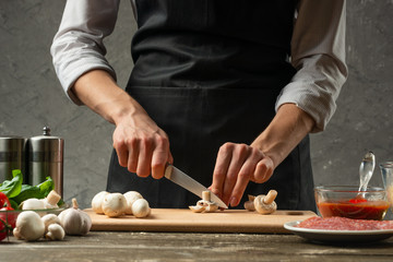 The concept of nutrition. The chef cuts mushrooms on the background of a concrete wall, with ingredients for cooking pizza, pizza sauce, for mushroom soup