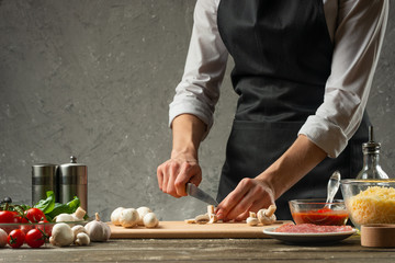 The concept of nutrition. The chef cuts mushrooms on the background of a concrete wall, with the ingredients for the preparation of pizza, pizza sauce, With empty space