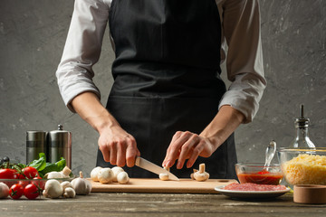 The concept of nutrition. The chef cuts mushrooms on the background of a concrete wall, with ingredients for cooking pizza, pizza sauce, for mushroom soup