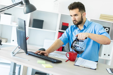 A young man stands in the office at the computer table and brews his coffee.