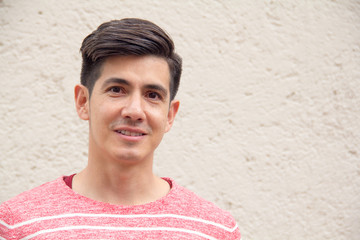 An attractive handsome young smiling Latino man with a trendy modern hair style in a striped red sweater looks direct to camera against a cream wall. With room for text / copy.