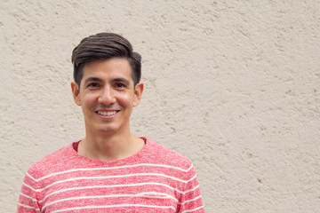 An attractive handsome young smiling Latino man with a trendy modern hair style in a striped red sweater looks direct to camera against a cream wall. With room for text / copy.