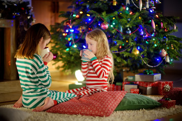 Two cute happy girls having hot chocolate by a fireplace in a cozy dark living room on Christmas eve.