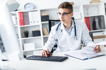 A young man in a white robe sitting at a table in the office. He holds a pen in his hand and works with documents and a computer.