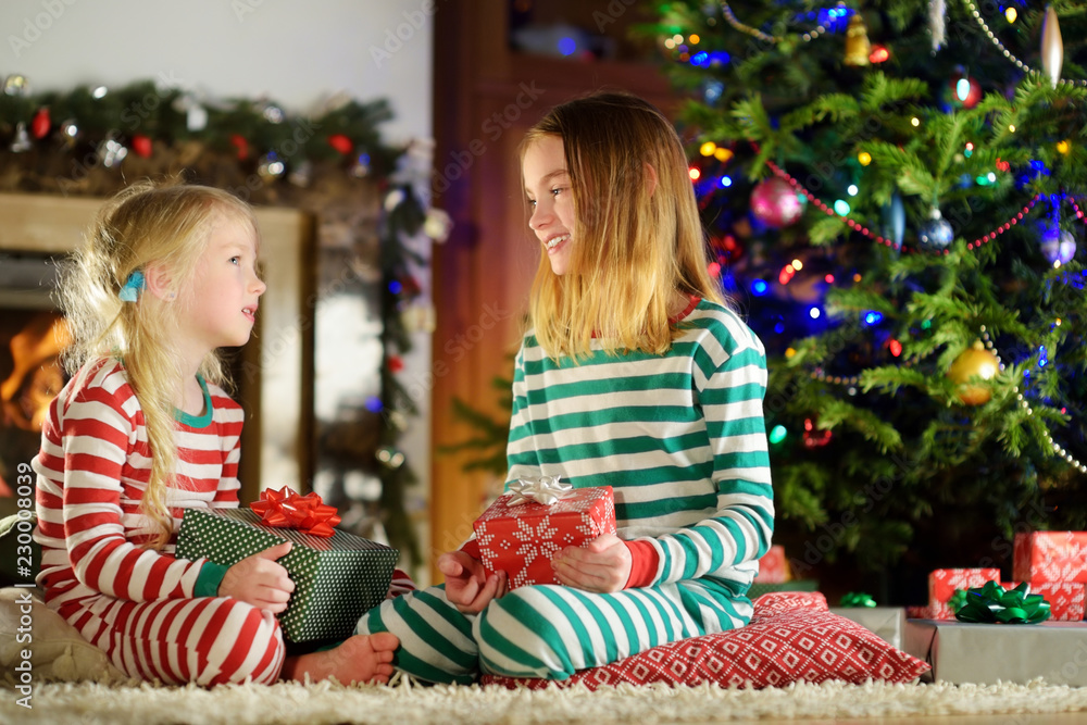 Wall mural happy little sisters wearing christmas pajamas opening gift boxes by a fireplace in a cozy dark livi