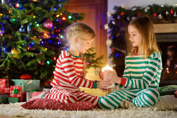 Fototapeta na wymiar Happy little girls wearing Christmas pajamas playing by a fireplace in a cozy dark living room on Christmas eve.
