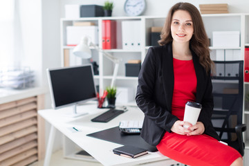 A young girl sat down on a table in the office and holding a glass of coffee.