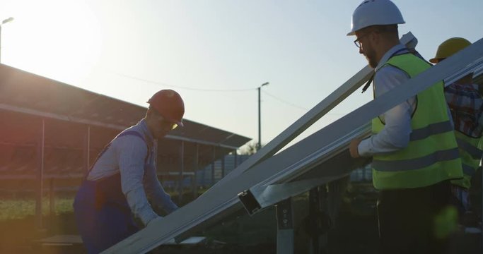 Medium shot of three workers in a uniform and hardhat installing photovoltaic panels on a metal basis on a solar farm