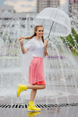 Young pretty girl with two braids in yellow boots and with transparent umbrella stands near fountain.