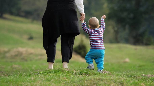 Baby child holding mother hand walking on green nature, kid learn to walk 