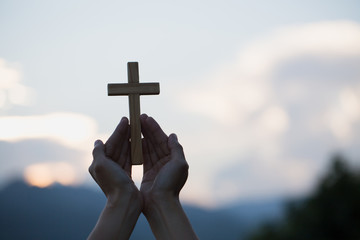 Woman hands holding holy lift of christian cross with light sunset background.