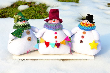 Three toys of small snowman in colorful hats and scarves are standing on the snow.