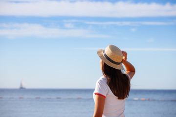 Fototapeta na wymiar Beautiful girl looks at the sea. Young girl in a hat looking at a calm sea and blue skies back view.