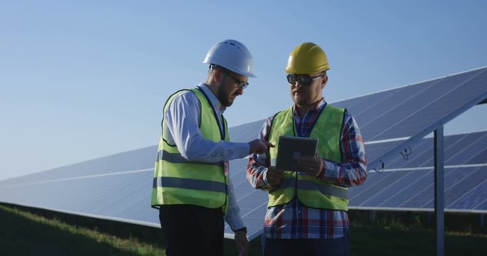 Medium shot of two electrical workers reviewing documents on a tablet during an inspection inbetween long rows of photovoltaic solar panels