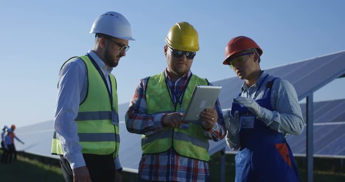 Medium shot of three electrical workers reviewing documents on a tablet during an inspection inbetween long rows of photovoltaic solar panels