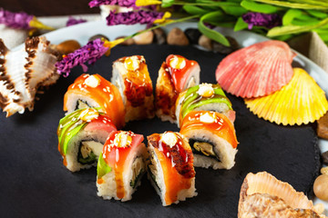 Rainbow sushi rolls decorated with teriyaki sauce at stone tray background served with lavender flowers and seashells.