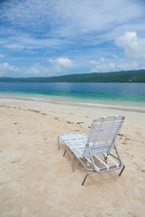 Fototapeta na wymiar Small plastic deck chair standing on sand of Cayo Levantado beach against beautiful water of Atlantic Ocean in the Samana Bay. Lounge is white and lightwaight. water is blue and sky has some clouds.