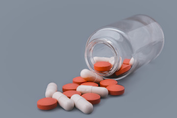 White and orange pills are scattered from a glass bottle on a gray background.