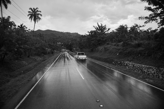 monochrome reportage photo of road going from mountain on Samana island in Dominican Republic. Sky is heavy and cloudy. A lot of trees are growing near the road. White pick-up is on the photo