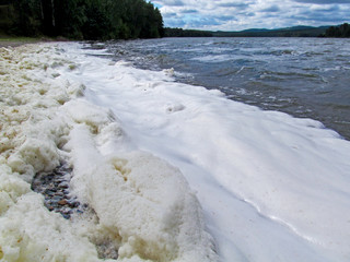 A lot of foam along the shore created by waves of water, close-up view. Summer scene of a lake or river. Concept of pollution of nature and a ecology problems of the environment.