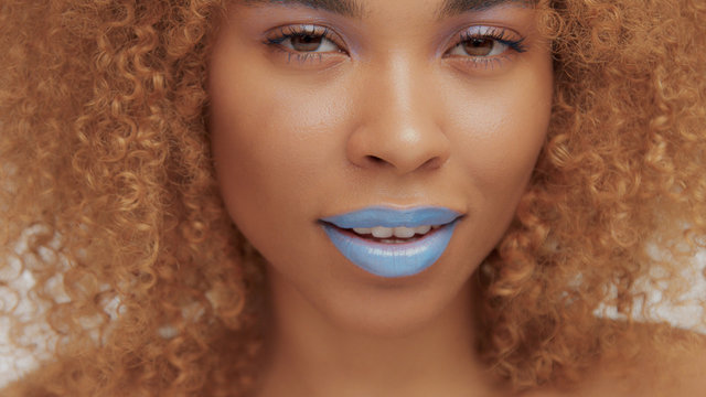mixed race black blonde model with curly hair closeup of mouth painted with bright blue lipstick smiling and watching to the camera