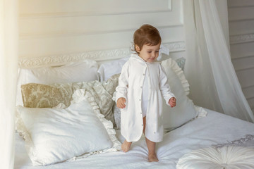 Funny and cute brunette little smiling girl playing jumping on bed in light bedroom. White interior with big bed. Childhood, preschool, youth, relax concept