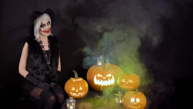 Scary beautiful girl witch celebrates halloween with funny glowing burning pumpkins in smoke. Female sorceress with gray hair scares, conjures, casts a spell