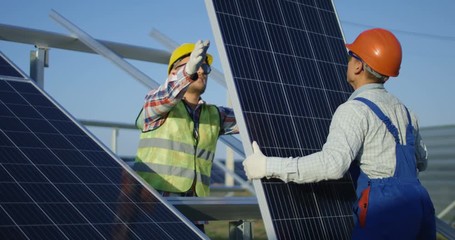 Medium shot of two workers in safety vests and hardhats installing photovoltaic panels on a metal basis at a solar farm