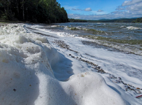 View of the lake from the beach, covered with a large surface of white foam. Unusual summer water landscape. Concept of pollution of nature and a ecology problems of the environment.
