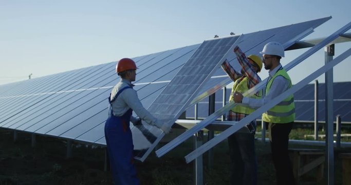 Medium shot of three workers in a uniform and hardhat installing photovoltaic panels on a metal basis on a solar farm