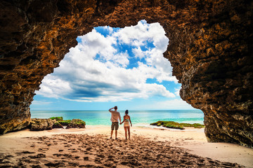 A loving couple enjoying the breathtaking views of the tropical sandy beach and sea from mountain cave - obrazy, fototapety, plakaty