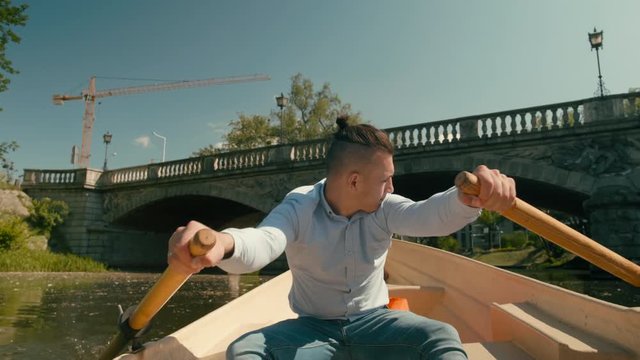 Young Millennial Man in a Blue Shirt is Paddling and Sculling Boat on the City River in Summer