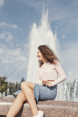 Vertical shot of hot pinup woman in trendy pink top and denim skirt, sitting on granite border of fountain, laughing out loud, having good time. Authentic vivid portrait of Caucasian female outdoors.
