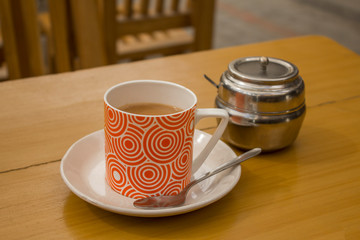 milk tea drink in a red mug on a saucer with a spoon on a wooden table with a metal sugar bowl