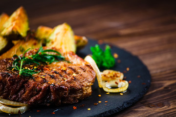 Grilled steak with baked potatoes and vegetables served on black stone on wooden table