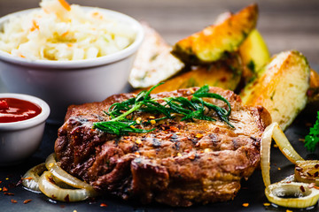 Grilled steak with baked potatoes and vegetables served on black stone on wooden table