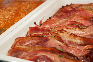 Closeup shot of trays of sliced grilled bacon and baked beans, healthy cooked breakfast options, adding protein and fiber to a diet. Horizontal shot with selective focus, concept for buffet-style meal