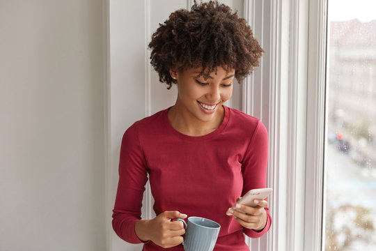 Happy pretty black girl happy to develop her own blog, rejoices having many followers, reads comments, drinks hot beverage, dressed in red clothes, stands near window. Hipster dials phone number