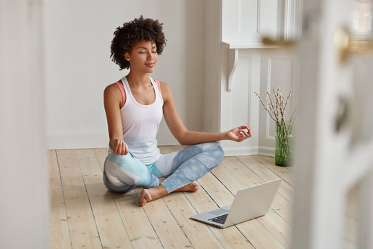Relaxed dark skinned woman with sporty body, sits in zen pose, keeps legs crossed, watches yoga lessons on laptop computer using internet, meditates on floor in empty room. Wellness concept.